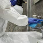 Close-up of a person disinfecting a bathroom sink with gloves and spray bottle.