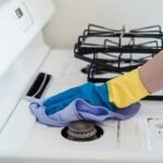 A close-up of a hand in gloves cleaning a gas stove with a cloth.