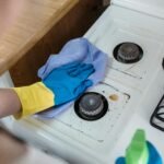 A person wearing rubber gloves cleaning a dirty gas stove with a cloth and spray bottle.