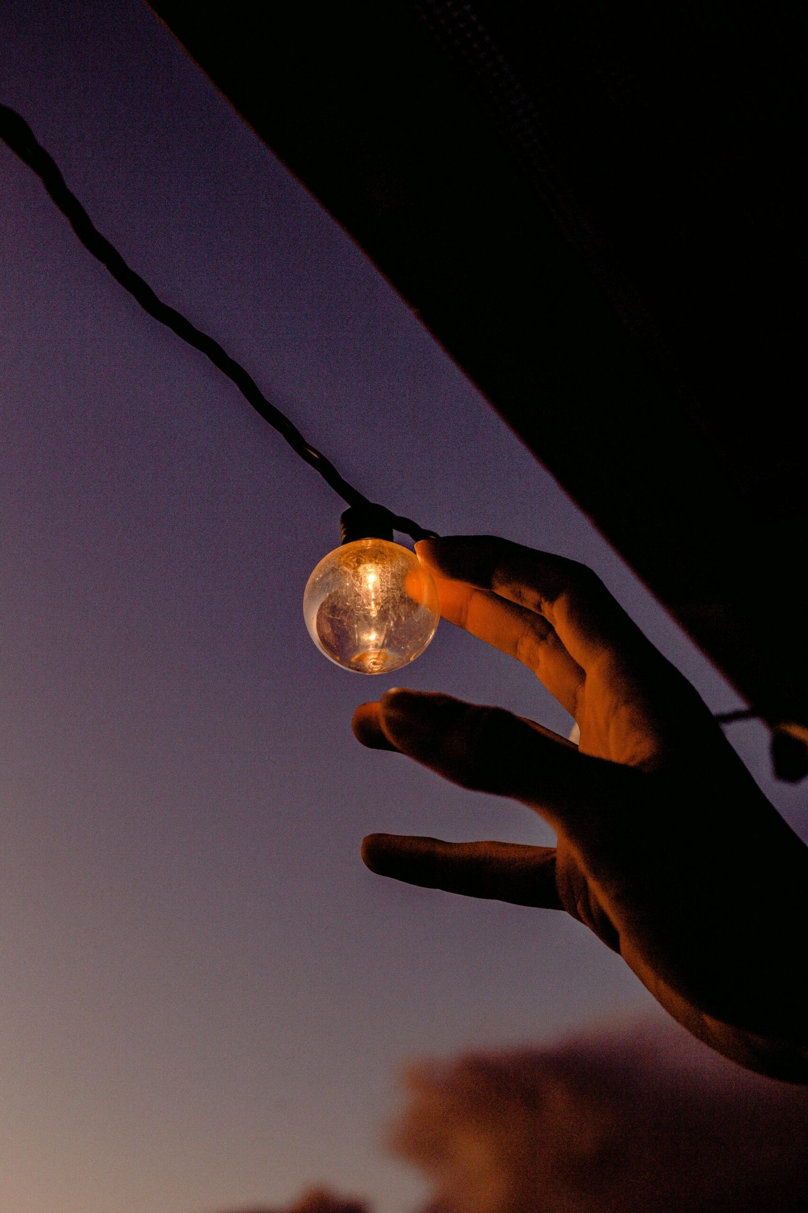 Silhouette of a hand touching a glowing string light bulb against a dramatic sunset sky.
